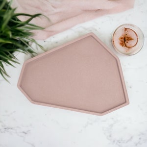 May include: A light pink, irregularly shaped, concrete tray with a speckled surface. The tray is on a white marble background.