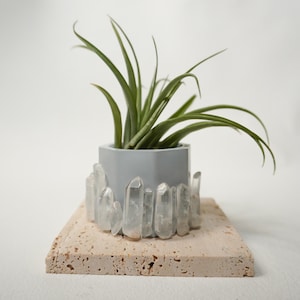 May include: A hexagonal gray concrete planter adorned with clear, vertical quartz crystals around its base. A green air plant with long, slender, pointed leaves sits inside the container, which rests on a square, porous, beige travertine tile.