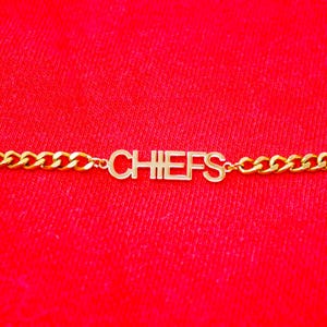 Könnte beinhalten: Ein goldenes Kettenarmband mit dem Wort "CHIEFS" in Blockbuchstaben.