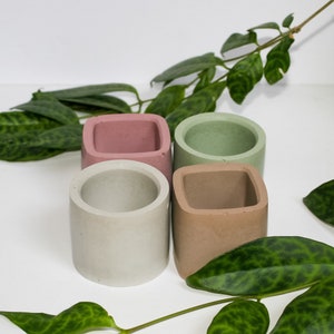 May include: Four small concrete planters in different colours and shapes. The planters are arranged in a square formation. The planters are light pink, light green, light grey, and light brown.