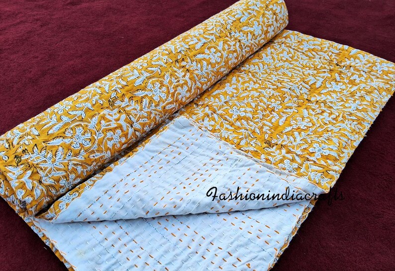Puede incluir: Una colcha kantha amarilla y blanca con un patr&oacute;n floral. La colcha est&aacute; doblada por la mitad y tiene un respaldo blanco con costuras amarillas.