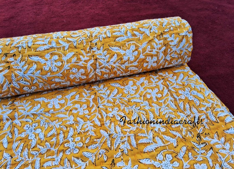 Puede incluir: Una colcha kantha amarilla con un patr&oacute;n floral blanco. La colcha est&aacute; doblada por la mitad y la tela es texturizada.