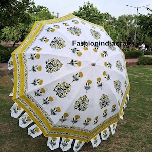 May include: A white parasol featuring a repeating pattern of yellow marigold flowers with green stems and leaves. The edge of the parasol is trimmed with a yellow border and small, dangling white fabric tabs. The text FashionIndiacrafts is visible.