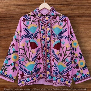 Peut inclure: Veste rose ornée de broderies florales bleues, jaunes et bordeaux. La veste a un col montant et des manches longues. Le texte "Fashionindiacrafts" est visible. Suspendue à un cintre en bois.