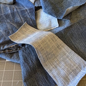 May include: A collection of denim fabric scraps in various shades of blue, including light blue, medium blue, and dark blue. The fabric is arranged in a pile, with some pieces overlapping.