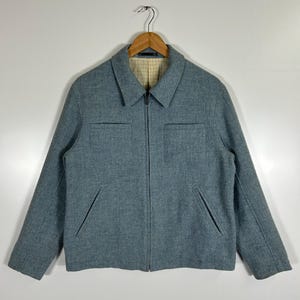 Könnte beinhalten: Hellblaue Jacke mit durchgehendem Reißverschluss, zwei Brusttaschen und zwei Seitentaschen. Die Jacke hat einen Kragen und ein kariertes Futter. Sie hängt an einem Holzbügel vor weißem Hintergrund.