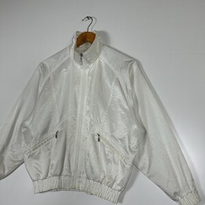 May include: A white, vintage-style jacket with a high collar and a full-length zipper. The jacket features two front pockets with zippers and a ribbed hem and cuffs. The material has a subtle, textured pattern. The jacket is hanging on a wooden hanger.