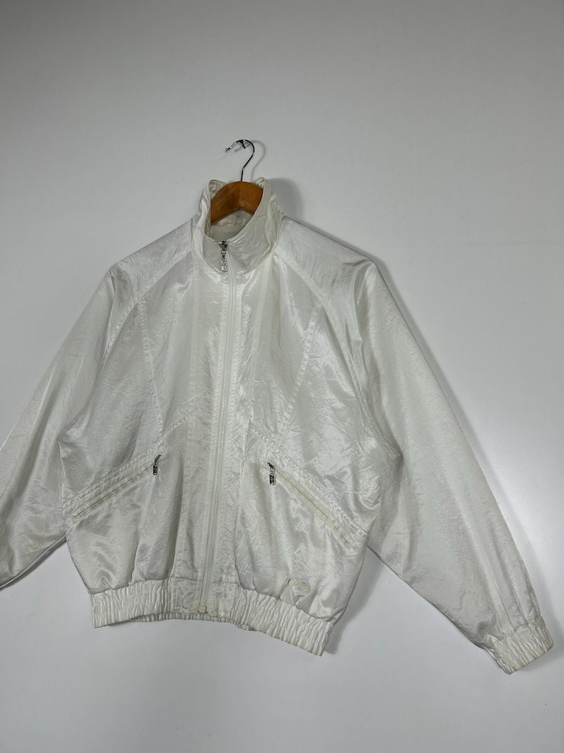 May include: A white, vintage-style jacket with a high collar and a full-length zipper. The jacket features two front pockets with zippers and a ribbed hem and cuffs. The material has a subtle, textured pattern. The jacket is hanging on a wooden hanger.