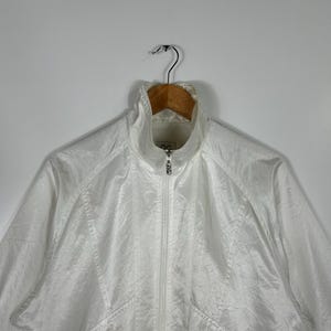 May include: White athletic jacket with a high collar and a full-length zipper. The jacket has two front pockets with zippers and is hanging on a wooden hanger. The material has a slight sheen.