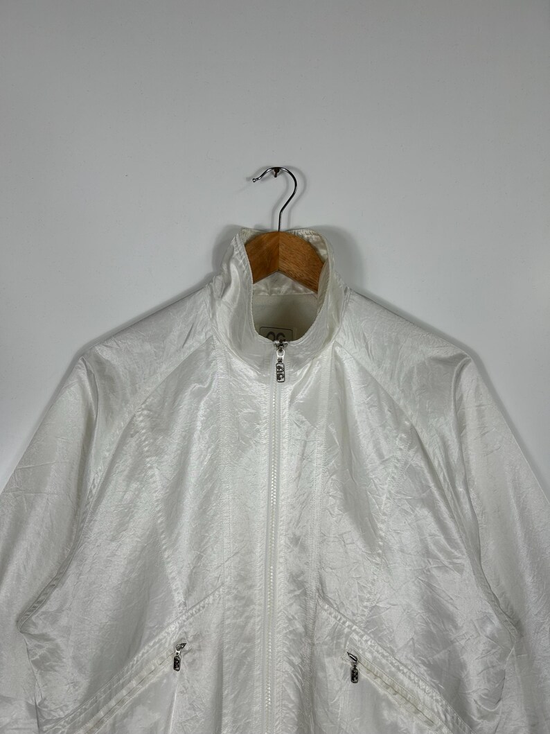 May include: White athletic jacket with a high collar and a full-length zipper. The jacket has two front pockets with zippers and is hanging on a wooden hanger. The material has a slight sheen.