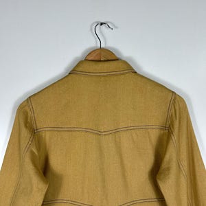 Peut inclure: Une veste &agrave; manches longues jaune moutarde dot&eacute;e d'un col classique et de surpiq&ucirc;res contrastantes marron fonc&eacute;. Le v&ecirc;tement pr&eacute;sente des coutures horizontales sur la poitrine et le dos, des manches longues, et est sur un cintre en bois.