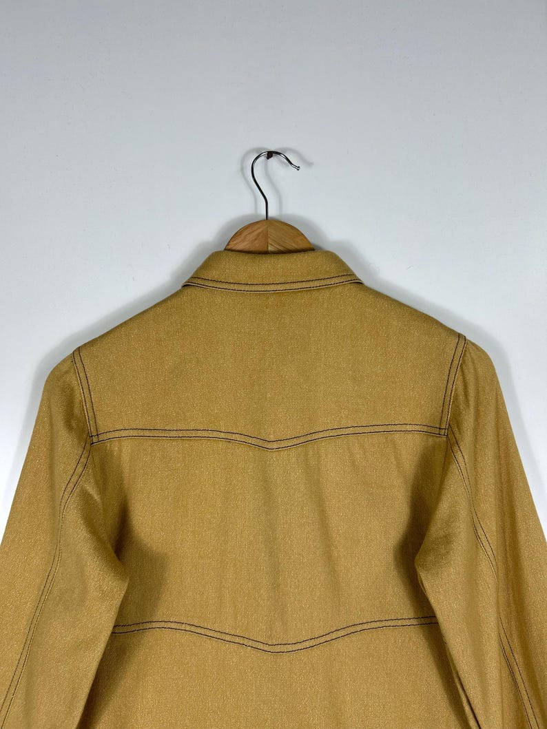 Peut inclure: Une veste &agrave; manches longues jaune moutarde dot&eacute;e d'un col classique et de surpiq&ucirc;res contrastantes marron fonc&eacute;. Le v&ecirc;tement pr&eacute;sente des coutures horizontales sur la poitrine et le dos, des manches longues, et est sur un cintre en bois.