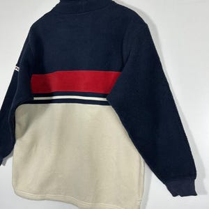 May include: A navy blue, red, and cream-colored fleece pullover. The top portion is navy blue with a red stripe and white accent. The bottom half is cream-colored. The pullover has a high collar and long sleeves.