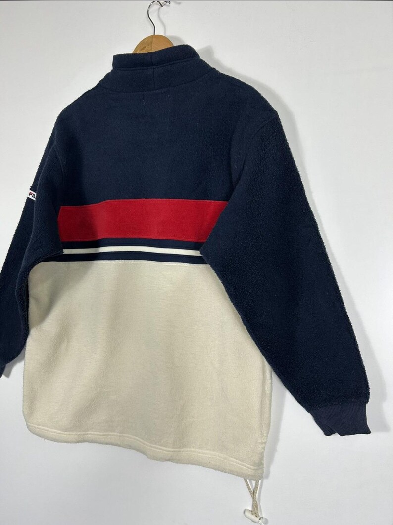 May include: A navy blue, red, and cream-colored fleece pullover. The top portion is navy blue with a red stripe and white accent. The bottom half is cream-colored. The pullover has a high collar and long sleeves.
