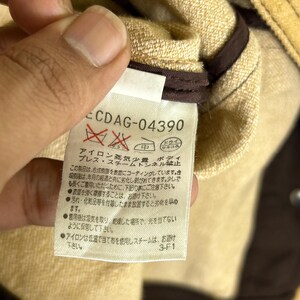 Peut inclure: Une &eacute;tiquette d'entretien de v&ecirc;tement blanche avec du texte et des symboles noirs, portant le code CDAG-04390. L'&eacute;tiquette contient des instructions en japonais concernant le repassage, la vapeur et le stockage, avec un bord en tissu marron.