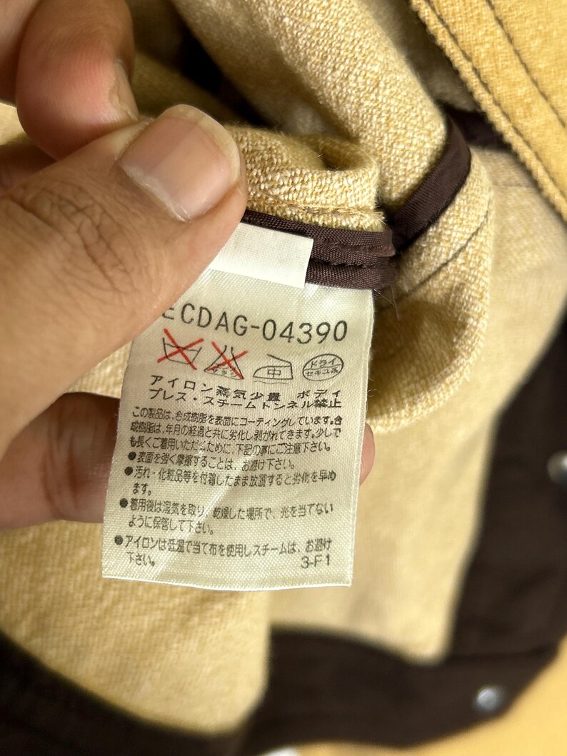 Peut inclure: Une &eacute;tiquette d'entretien de v&ecirc;tement blanche avec du texte et des symboles noirs, portant le code CDAG-04390. L'&eacute;tiquette contient des instructions en japonais concernant le repassage, la vapeur et le stockage, avec un bord en tissu marron.