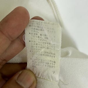 May include: Close-up of a white fabric tag with Japanese text. The tag is attached to a white garment. The text provides care instructions, including washing and drying recommendations.