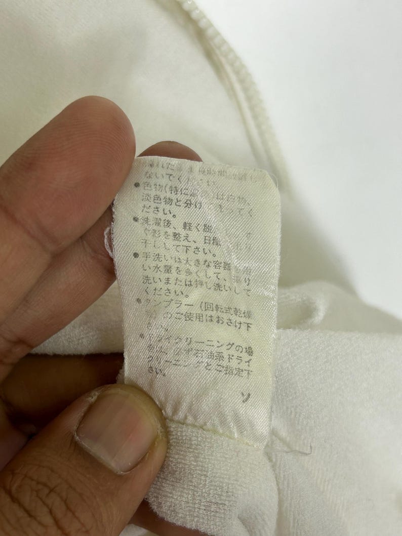 May include: Close-up of a white fabric tag with Japanese text. The tag is attached to a white garment. The text provides care instructions, including washing and drying recommendations.