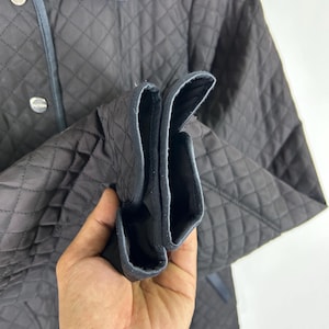 May include: A dark brown quilted jacket with a diamond pattern. The jacket has a collar with a blue trim and silver buttons. The sleeves are cuffed, revealing a black lining. The jacket is made of a soft, quilted material.