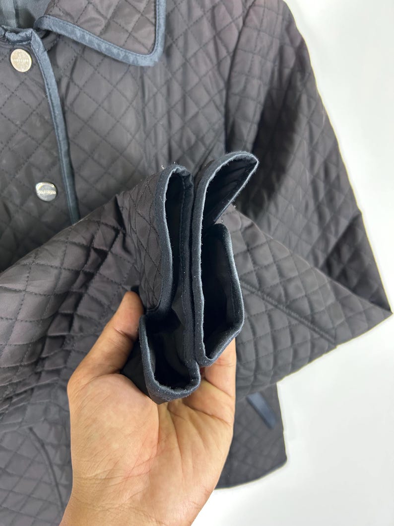 May include: A dark brown quilted jacket with a diamond pattern. The jacket has a collar with a blue trim and silver buttons. The sleeves are cuffed, revealing a black lining. The jacket is made of a soft, quilted material.