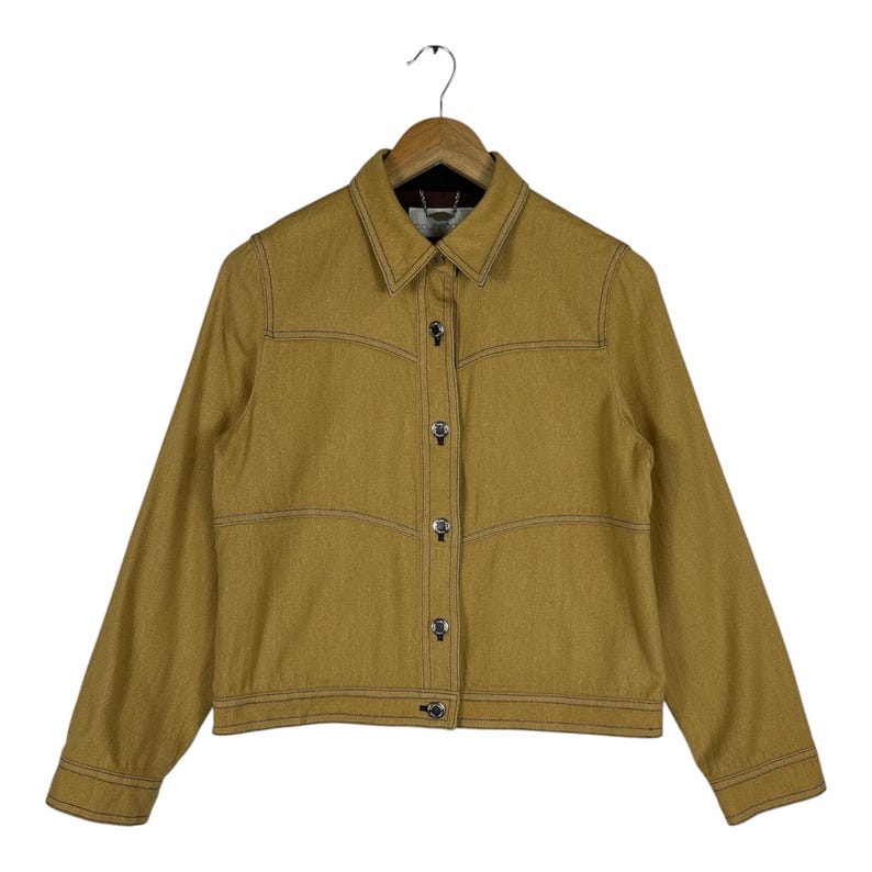 Peut inclure: Une veste &agrave; manches longues jaune moutarde dot&eacute;e d'un col classique, d'une fermeture boutonn&eacute;e sur le devant avec des boutons m&eacute;talliques et de coutures horizontales d&eacute;coratives sur la poitrine. Le v&ecirc;tement est pr&eacute;sent&eacute; sur un cintre en bois.