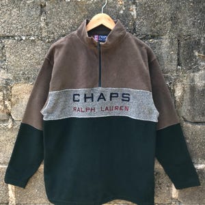 May include: A color-blocked fleece pullover in brown, gray, and dark green. The top features a half-zip closure and the brand name "CHAPS RALPH LAUREN" across the chest in navy and red lettering. The garment is hanging on a wooden hanger.