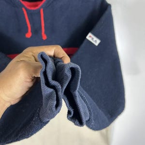 May include: Navy blue Fila sweatshirt with red accents. The sweatshirt features a red collar and a red stripe across the chest. The Fila logo is visible on the chest and sleeve. The bottom portion of the sweatshirt is off-white.