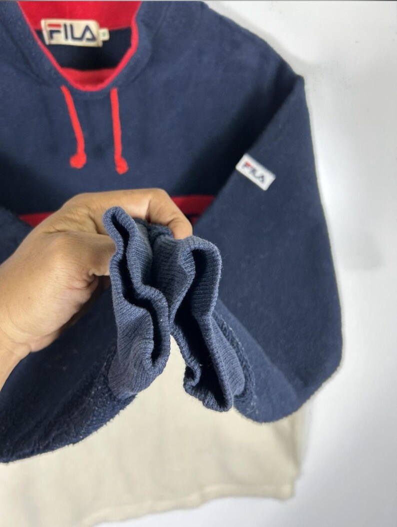 May include: Navy blue Fila sweatshirt with red accents. The sweatshirt features a red collar and a red stripe across the chest. The Fila logo is visible on the chest and sleeve. The bottom portion of the sweatshirt is off-white.