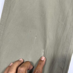 Puede incluir: Primer plano de unos pantalones de color caqui. La tela es de un tono liso y neutro con costuras visibles a lo largo de las costuras. Hay peque&ntilde;as manchas oscuras en la tela. Una mano se&ntilde;ala los pantalones.