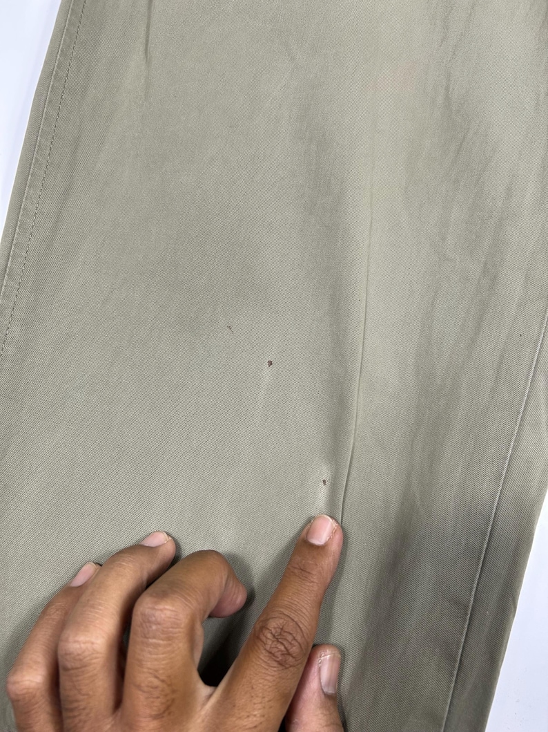 Puede incluir: Primer plano de unos pantalones de color caqui. La tela es de un tono liso y neutro con costuras visibles a lo largo de las costuras. Hay peque&ntilde;as manchas oscuras en la tela. Una mano se&ntilde;ala los pantalones.