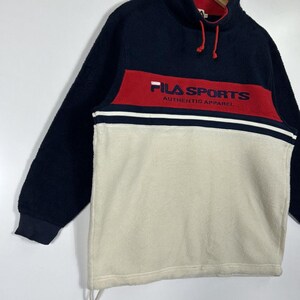 May include: A navy blue, red, and cream-colored fleece pullover. The top section is navy blue, with a red band across the chest featuring "FILA SPORTS" in white. The lower section is cream-colored. The pullover has a high collar and drawstring.