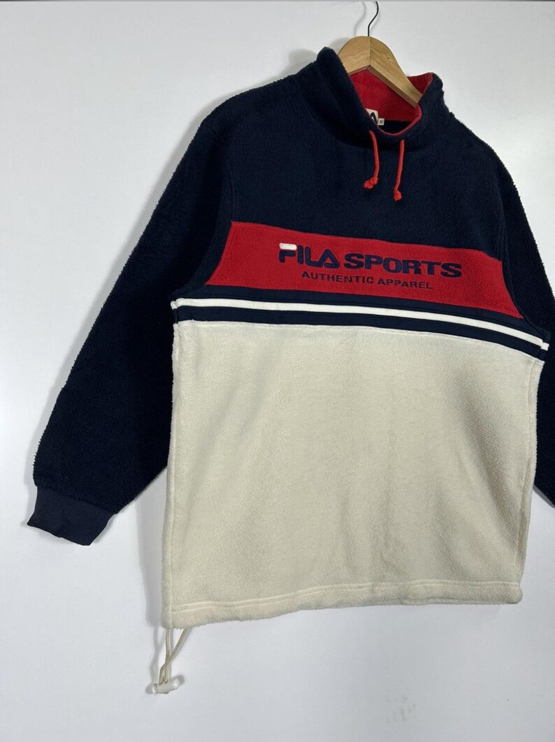 May include: A navy blue, red, and cream-colored fleece pullover. The top section is navy blue, with a red band across the chest featuring "FILA SPORTS" in white. The lower section is cream-colored. The pullover has a high collar and drawstring.