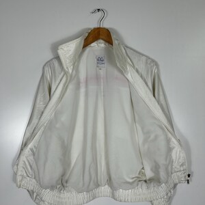 May include: A white vintage track jacket with a high collar and a full zipper. The jacket has a shiny, slightly textured appearance and a ribbed hem and cuffs. The interior is white, and the jacket is hanging on a wooden hanger.
