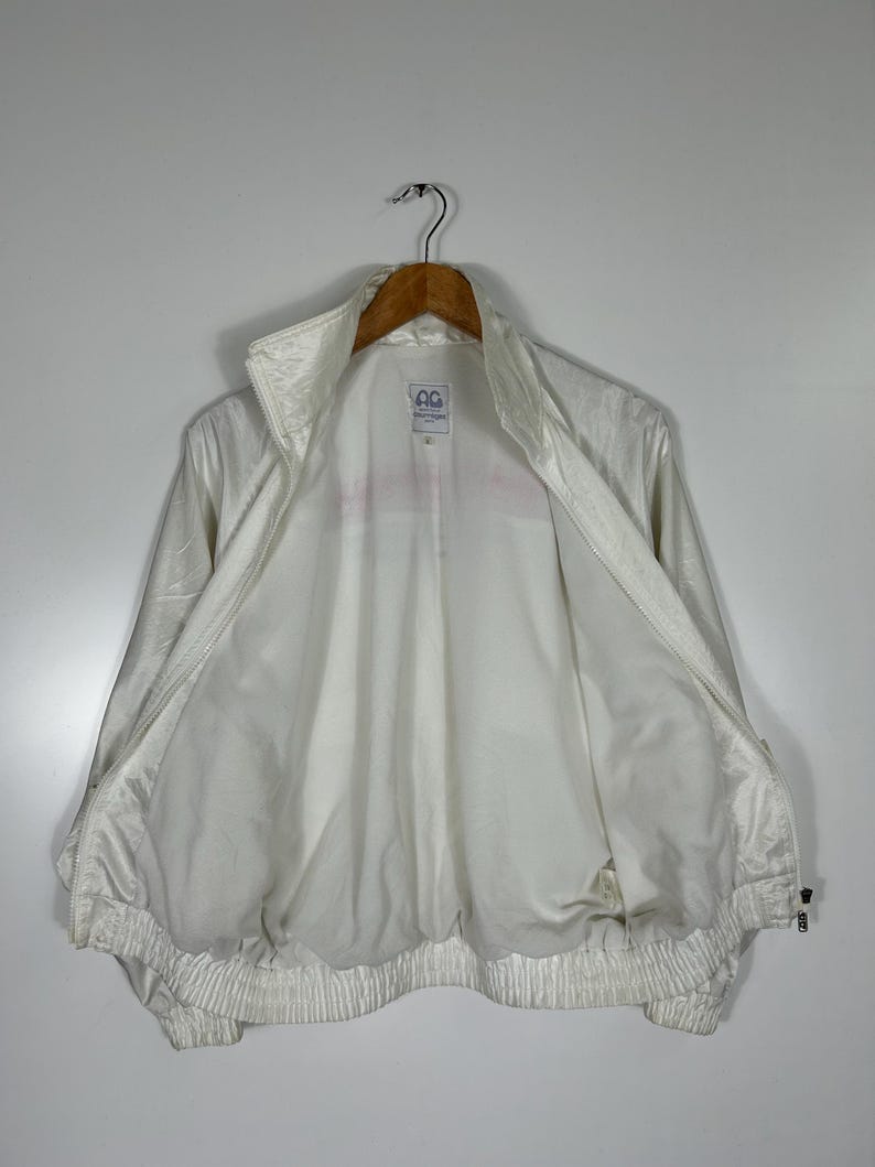 May include: A white vintage track jacket with a high collar and a full zipper. The jacket has a shiny, slightly textured appearance and a ribbed hem and cuffs. The interior is white, and the jacket is hanging on a wooden hanger.