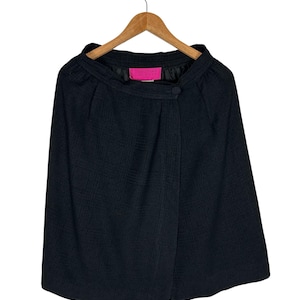 Puede incluir: Una chaqueta negra estilo capa con un patrón texturizado, colgada de una percha de madera. La chaqueta tiene un cierre de botón y una etiqueta rosa con texto. La prenda es de color oscuro y parece estar hecha de una tela tejida.