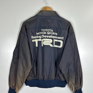 May include: A navy blue jacket featuring the text "TOYOTA MOTOR SPORTS Racing Development TRD" in white on the back. The jacket has a dark blue ribbed collar and cuffs. It is suspended from a wooden hanger.
