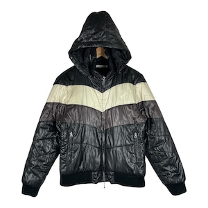 Könnte beinhalten: Eine schwarze Kapuzen-Steppjacke mit einem horizontalen, cremefarbenen Chevron-Streifen über der Brust und einem dunkelgrauen Band darunter. Die Jacke verfügt über einen durchgehenden Frontreißverschluss, zwei seitliche Reißverschlusstaschen sowie schwarze Rippbündchen an Ärmeln und Saum.