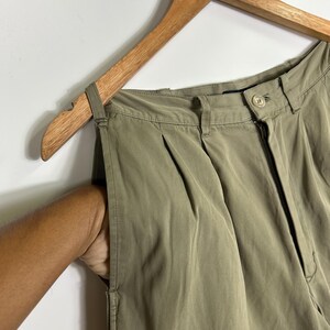 Puede incluir: Pantalones plisados ​​verde oliva colgando de una percha de madera. Los pantalones tienen cierre de cremallera y bot&oacute;n. Una mano est&aacute; en el bolsillo de los pantalones.