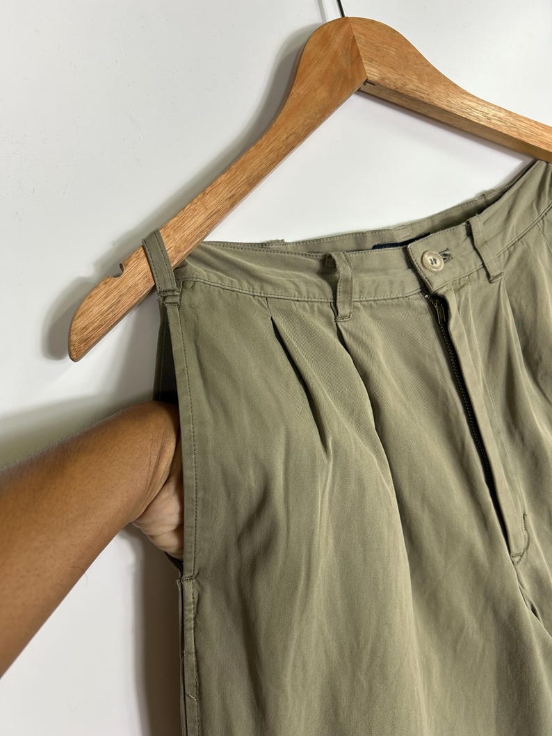 Puede incluir: Pantalones plisados ​​verde oliva colgando de una percha de madera. Los pantalones tienen cierre de cremallera y bot&oacute;n. Una mano est&aacute; en el bolsillo de los pantalones.