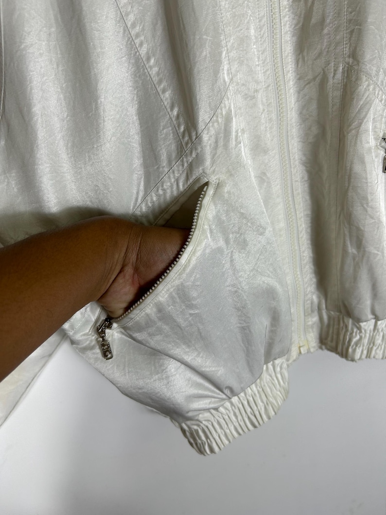 May include: Off-white, vintage windbreaker jacket with a full zipper closure and two front pockets. The jacket has a ribbed hem and cuffs. The zipper pull has a decorative design. The fabric has a subtle sheen.
