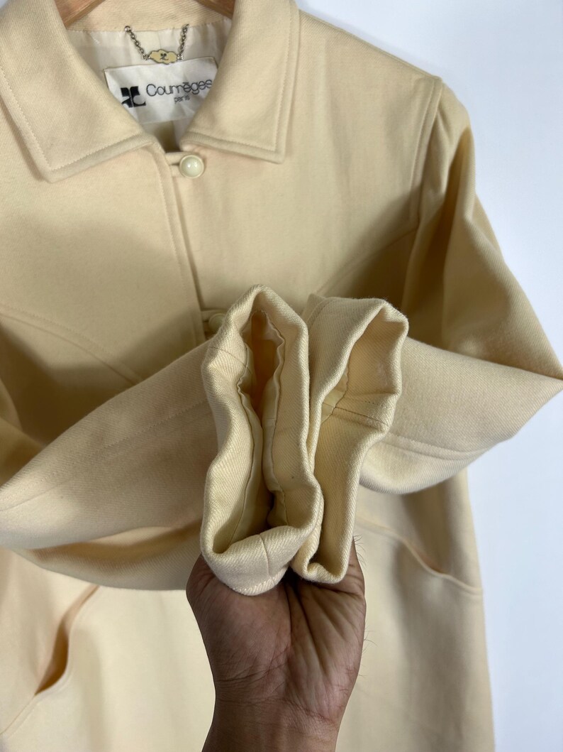 May include: A cream-coloured jacket with a pointed collar, long sleeves, and round buttons. The interior label reads Courr&egrave;ges Paris. The garment features a structured design with visible seams and a smooth, solid-coloured fabric construction.