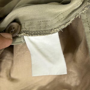 Puede incluir: Primer plano del interior de una prenda beige, mostrando un bot&oacute;n, costuras y una etiqueta blanca en blanco. La tela parece ser de algod&oacute;n o mezcla de peso medio, con una textura suave. La etiqueta es rectangular.