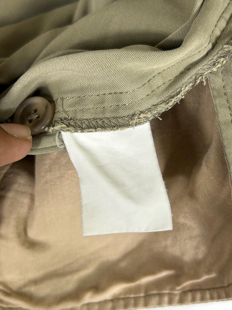 Puede incluir: Primer plano del interior de una prenda beige, mostrando un bot&oacute;n, costuras y una etiqueta blanca en blanco. La tela parece ser de algod&oacute;n o mezcla de peso medio, con una textura suave. La etiqueta es rectangular.