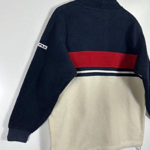 May include: A navy blue, red, and cream-colored fleece pullover. The top half is navy blue, with a red stripe and white stripe across the chest. The bottom half is cream-colored. The Fila logo is on the left sleeve.