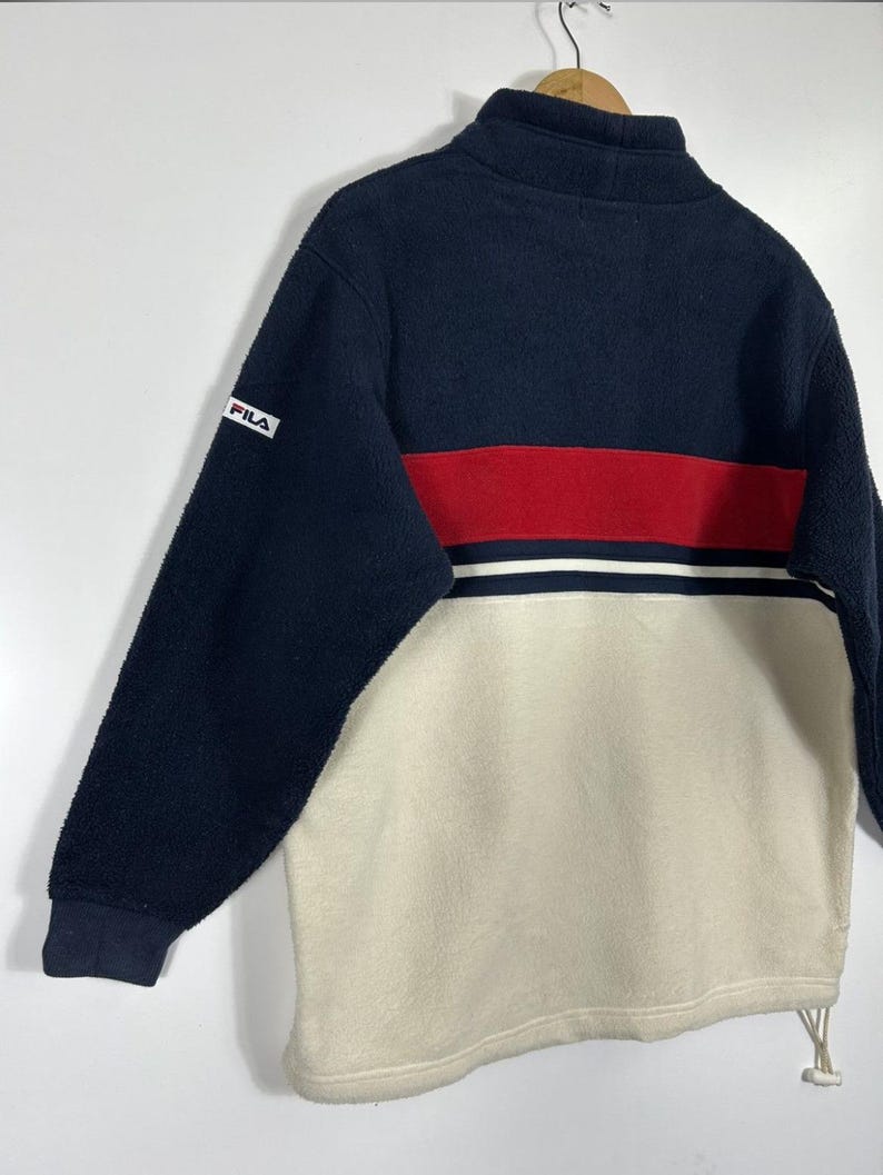 May include: A navy blue, red, and cream-colored fleece pullover. The top half is navy blue, with a red stripe and white stripe across the chest. The bottom half is cream-colored. The Fila logo is on the left sleeve.