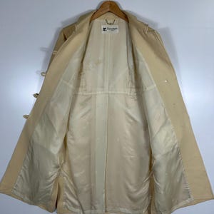 May include: A beige trench coat hanging on a wooden hanger, showing the interior lining. The coat features a light-coloured satin-like lining with a subtle repeating logo pattern and a white brand label at the neck that reads Courr&egrave;ges Paris.