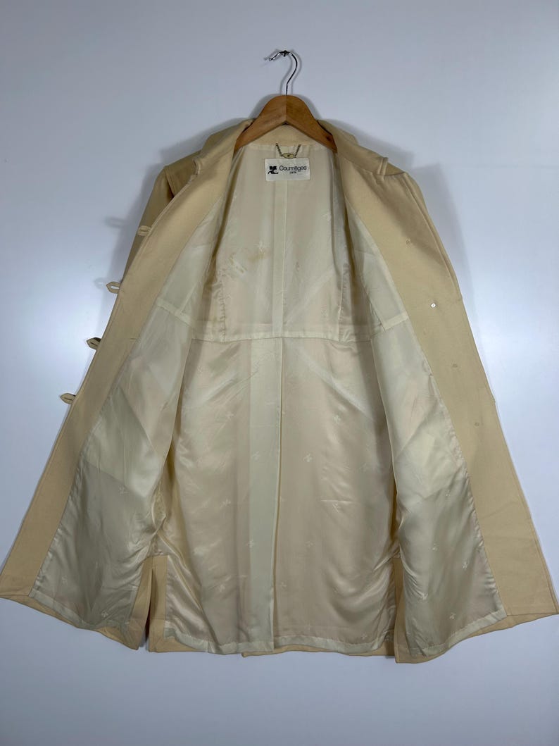 May include: A beige trench coat hanging on a wooden hanger, showing the interior lining. The coat features a light-coloured satin-like lining with a subtle repeating logo pattern and a white brand label at the neck that reads Courr&egrave;ges Paris.