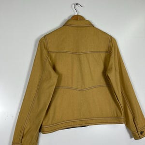Peut inclure: Une veste jaune moutarde dot&eacute;e d'un col classique, de manches longues avec poignets boutonn&eacute;s et de surpiq&ucirc;res contrastantes marron fonc&eacute;. Le panneau arri&egrave;re pr&eacute;sente des coutures horizontales avec un d&eacute;tail en forme de V, sur cintre.