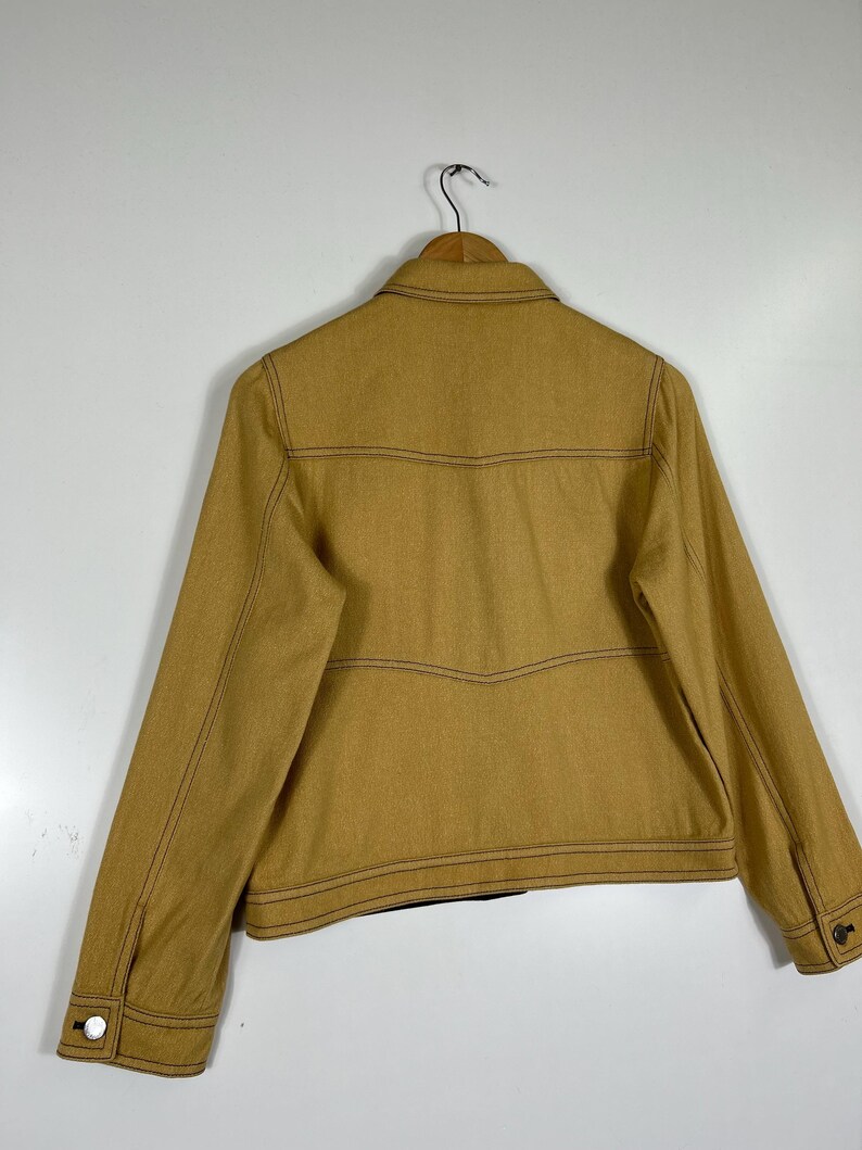 Peut inclure: Une veste jaune moutarde dot&eacute;e d'un col classique, de manches longues avec poignets boutonn&eacute;s et de surpiq&ucirc;res contrastantes marron fonc&eacute;. Le panneau arri&egrave;re pr&eacute;sente des coutures horizontales avec un d&eacute;tail en forme de V, sur cintre.