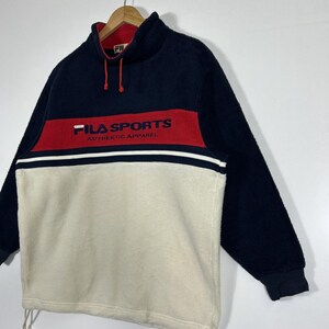 May include: A navy blue, red, and cream-colored FILA SPORTS fleece pullover. The pullover features a high collar, long sleeves, and the FILA SPORTS logo in red across the chest. The bottom half is cream-colored, with a white stripe separating it from the red band.